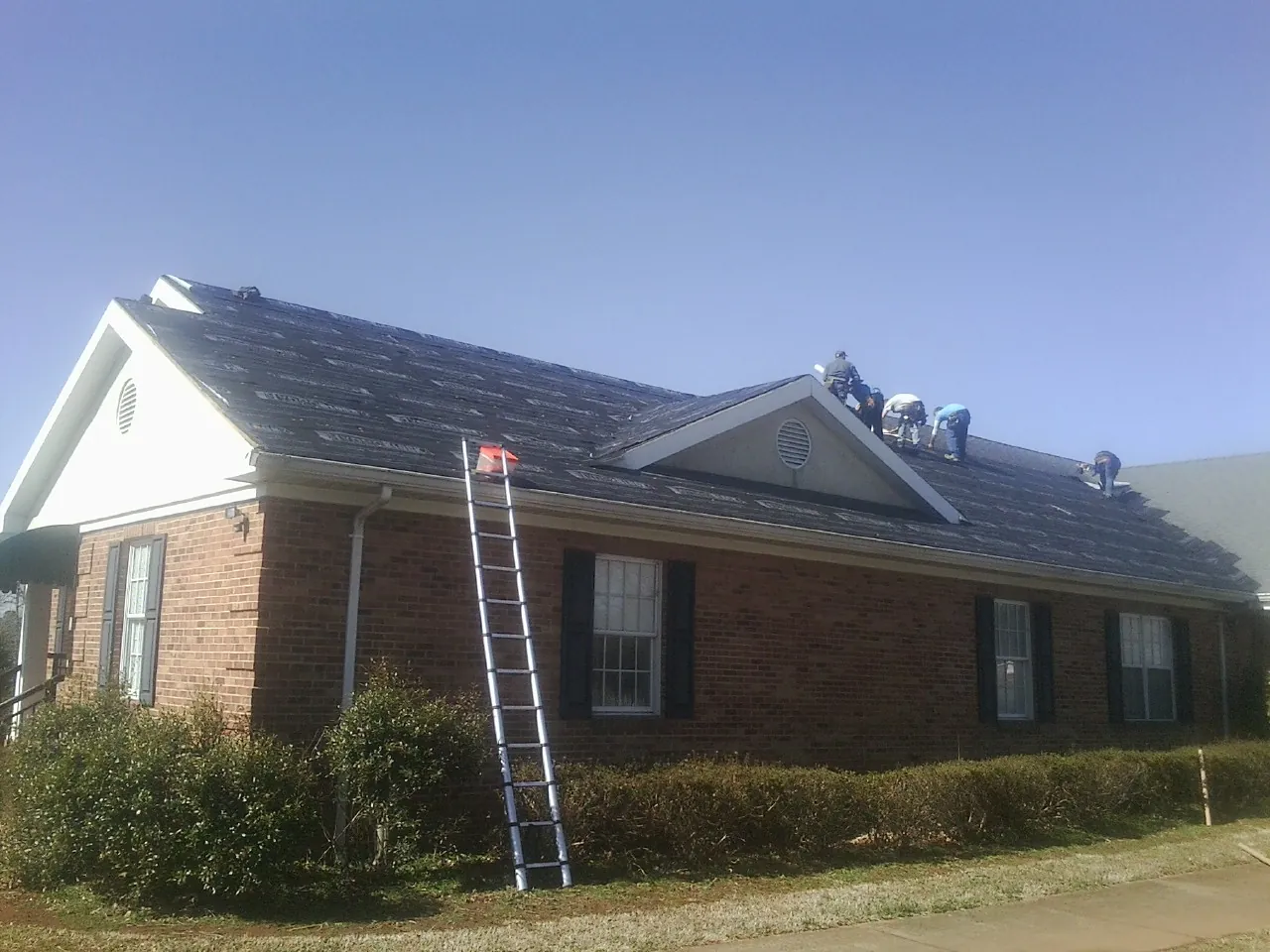 Crew tearing shingles off a brick ranch roof with a ladder set — Emergency Roof Repair work by our crew in Lanett