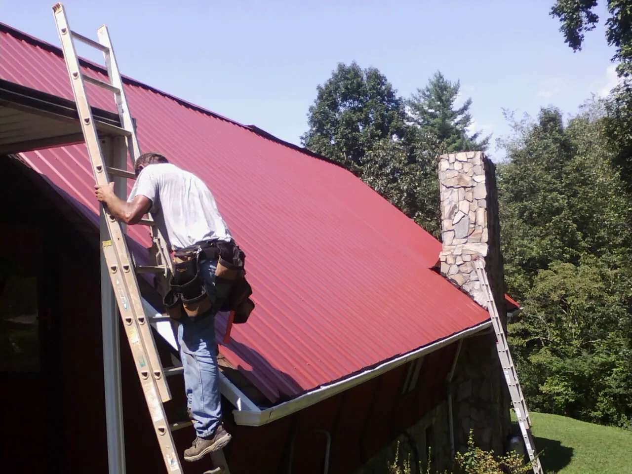 Roofer on a ladder beside a red metal roof and stone chimney — Soffit & Fascia Repair by Roofing Brothers in Lanett